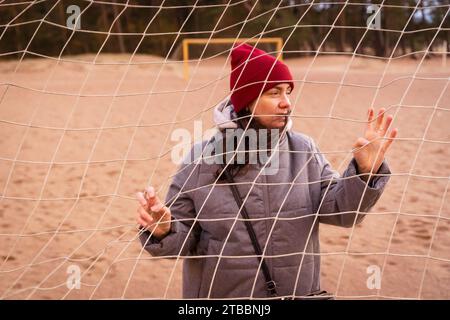 Giovane donna alla moda che indossa una giacca e un cappello autunnali e posa davanti a una rete da calcio sulla spiaggia. L'elegante donna è sicura di sé e Foto Stock