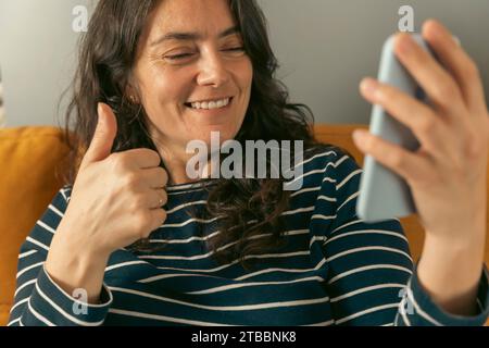 Donna di mezza età con i capelli castani sorridente mentre effettua una videochiamata sul suo smartphone e fa un gesto pollice su, mostrando la sua approvazione e la felicità Foto Stock