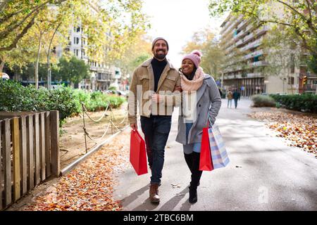 Felice coppia multirazziale che cammina in città per fare compere di natale insieme, tenendo in mano le borse regalo. Foto Stock