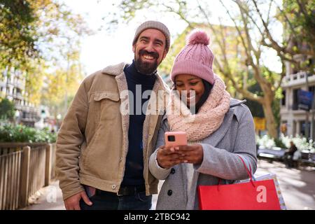 Felice coppia interrazziale che fa shopping natalizio, in piedi all'aperto usando insieme uno smartphone. Foto Stock