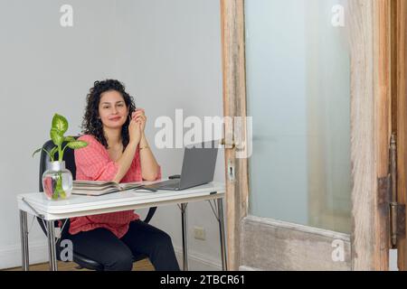 Giovane donna latina felice con i ricci, è seduta nel suo nuovo ufficio a casa, sorridendo e guardando la macchina fotografica, molto felice pronta a lavorare sulla sua avventura. Foto Stock