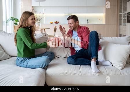 Moglie sorridente che tiene una confezione regalo con nastro rosso, congratulandosi con il marito per il compleanno e la data del matrimonio Foto Stock