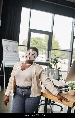 gioiosa donna d'affari afro-americana curvy che guarda la fotocamera vicino al luogo di lavoro in un ufficio moderno Foto Stock