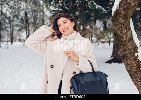 Giovane donna alla moda in posa all'aperto, indossa un cappotto bianco in pelliccia di pecora che tiene la borsa nera sotto le nevicate Foto Stock