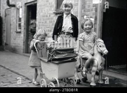 anni '1960, storia, bambini che giocano per strada fuori casa. In Gran Bretagna negli anni '50 e '60 i bambini piccoli nelle aree urbane giocherebbero liberamente fuori per strada senza supervisione. Qui vediamo una ragazza con la sua carrozza giocattolo costruita in carrozza, mentre sua sorella maggiore sul marciapiede è seduta sul suo cavallo dondolante. Foto Stock