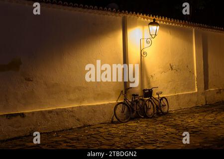 Paraty, Brasile. Biciclette sulla parete sotto la luce dei vecchi lampioni di notte. Strada fatta con pietre del periodo coloniale. Foto Stock