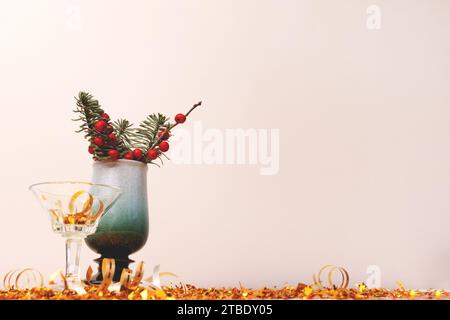 Bouquet di nobilis abete e rami di winterberry in vaso, piccolo vetro di cristallo e coriandoli d'oro davanti a sfondo beige. Foto Stock