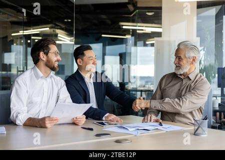 Tre uomini d'affari che hanno una riunione d'affari in ufficio, stringono la mano e sorridono, trattative di successo. Foto Stock