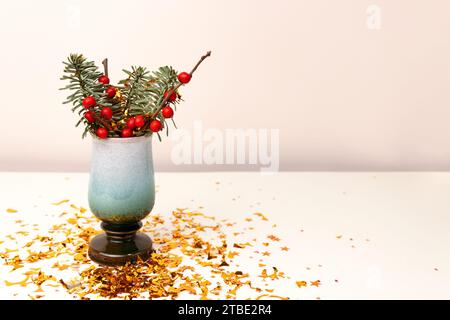 Bouquet di nobilis abete e rami di winterberry in vaso e coriandoli d'oro davanti a sfondo beige. Composizione festiva con spazio per il testo. Foto Stock