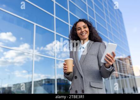 Felice donna d'affari latinoamericana passa davanti all'edificio di vetro, tiene una tazza di caffè al telefono, sorride e guarda la fotocamera. Foto Stock