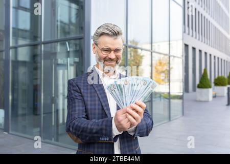 Un uomo d'affari incredibilmente felice ha in mano una pila di dollari, non riesce a credere alla sua fortuna, sorride e guarda i soldi. Foto Stock