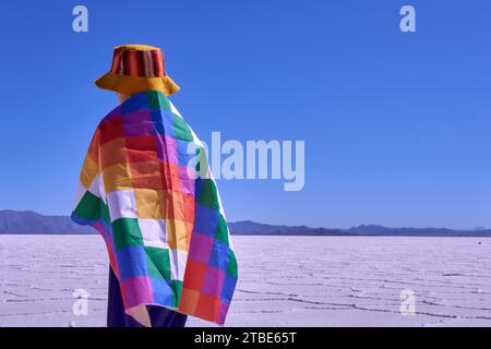 Ragazzo che cammina all'indietro con una bandiera di whipala sulla schiena e un cappello colorato attraverso le grandi distese saline, a Jujuy, Argentina. sullo sfondo il blu Foto Stock