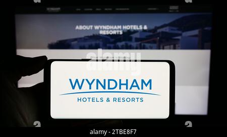 Persona che possiede il cellulare con il logo della società alberghiera statunitense Wyndham Hotels and Resorts Inc. Davanti alla pagina Web aziendale. Concentrarsi sul display del telefono. Foto Stock