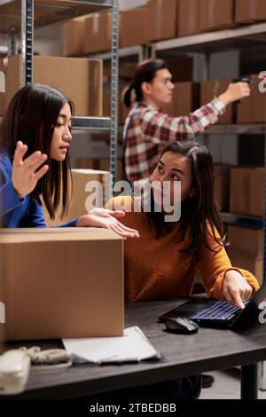 Operatore di magazzino che supervisiona il collega che esegue l'inventario sul laptop nel magazzino. Storehouse giovani dipendenti asiatiche che discutono della logistica delle merci e del programma di distribuzione Foto Stock