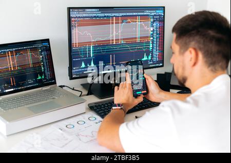 Il trader maschile siede in ufficio davanti a schermi con grafici e grafici finanziari, utilizzando smartphone, predice, analizza e studia la crescita o il declino della criptovaluta.Borsa, trading Foto Stock