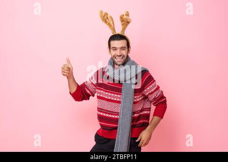 Lo studio natalizio ritrae un uomo caucasico sorridente che indossa maglione e un elegante cerchietto di renna con i pollici su uno sfondo isolato di colore rosa Foto Stock