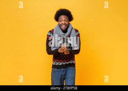 Un uomo afro-americano sorpreso e sorpreso che indossa maglione e sciarpa natalizi, girato in uno studio natalizio con sfondo isolato di colore giallo Foto Stock