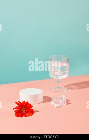 Un bicchiere di vino contenente acqua, un fiore rosso di gerbera, ghiaccio e un podio bianco rotondo spiccano su uno sfondo blu-rosa. Visualizza i prodotti su una temp Foto Stock