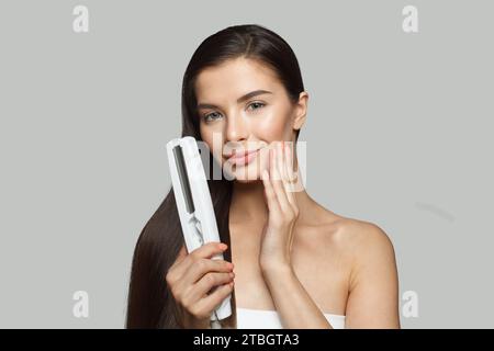 Donna attraente che usa ferro da stiro e liscia i suoi lunghi capelli castani su sfondo bianco, ritratto di bellezza da studio. Stiratura e acconciatura dei capelli Foto Stock