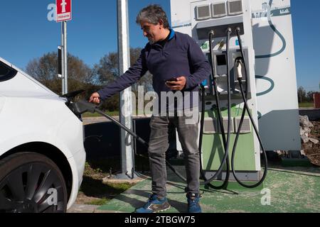 Uomo collega una spina CCS alla sua auto elettrica Tesla Model 3 a una stazione di ricarica rapida pubblica Foto Stock