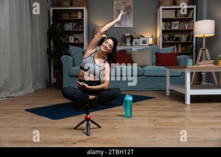 Giovane donna attraente che impara nuovi esercizi guardando esercitazioni di allenamento online tramite telefono a casa. Bella donna che pratica lezione di yoga avendo V Foto Stock