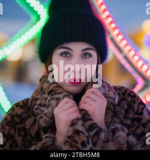 Ritratto di timida giovane donna con cappello nero e cappotto in pelliccia con motivo leopardato si erge sullo sfondo delle decorazioni delle luci di Natale e guarda la fotocamera Foto Stock