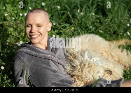 Una donna calva felice e contenta con un cane è sdraiata sull'erba verde in una giornata di sole. Concetto di gioia di vita. Domestico... Foto Stock