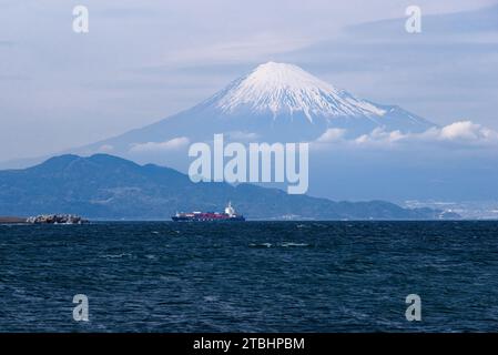 Questa funzione cattura la gloria e le dimensioni del Monte Fuji mentre incombe su una nave portacontainer che naviga verso il porto di Shimizu attraverso la baia di Suruga. Foto Stock