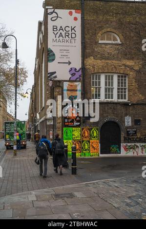 La gente passa accanto al Brick Shop all'angolo tra Back Yard Market e Brick Lane. Poster colorati sulla parete e indicazioni stradali per il mercato. Londra, Regno Unito Foto Stock