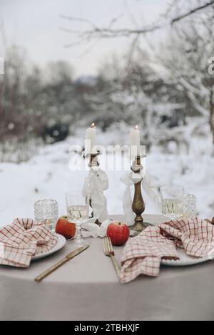 Tavolo natalizio all'aperto per due con bicchiere di vino, candele e tovaglioli a scacchi. Matrimonio invernale, San Valentino, festa di compleanno Foto Stock