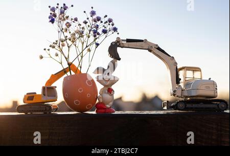 2 escavatori giocattolo, un uovo dipinto con fiori di gypsophila, un coniglio pasquale retroilluminato contro il cielo. Il concetto di Pasqua per la costruzione di compani Foto Stock