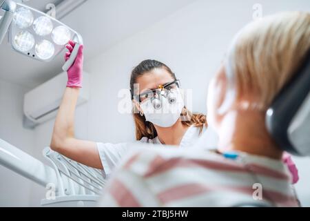 Appuntamento paziente clinica dentale in moderno reparto medico. Medico dentista in lenti d'ingrandimento che puntano la luce su giovane femmina pronta per la chirurgia dentaria. Foto Stock