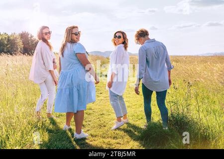 Quattro allegre donne sorridenti camminano lungo l'alta collina verde prato, ridendo e chiacchierando tra loro durante una passeggiata al tramonto. Amici della donna Foto Stock