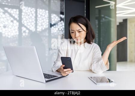Donna triste turbata sul posto di lavoro che tiene uno smartphone, donna d'affari depressa che naviga sui social media all'interno dell'ufficio, lavoratrice asiatica che usa un computer portatile al lavoro. Foto Stock