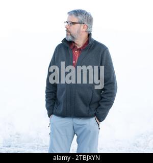 Uomo, 56 anni, capelli e barba grigi, in piedi all'aperto in inverno con neve sullo sfondo, mani in tasca, occhiali Foto Stock
