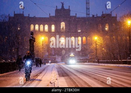 Monaco, Germania. 1 dicembre 2023. Spessi fiocchi di neve cadono dal cielo di fronte al Maximilianeum, sede del Parlamento di Stato bavarese, mentre un ciclista cavalca il Ponte Massimiliano innevato in primo piano. La statua di Pallade Atena è visibile sulla sinistra. Crediti: Matthias Balk/dpa/Alamy Live News Foto Stock