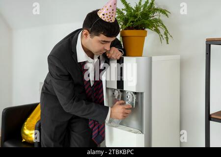 Uomo d'affari sbornico che versa acqua dal refrigeratore dopo la festa di Capodanno in ufficio Foto Stock