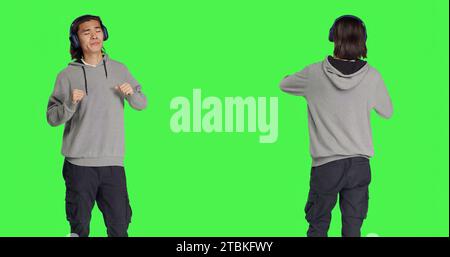 Il ragazzo positivo fa passi di danza funky sulla fotocamera, sentendosi groviglio mentre ascolta canzoni moderne. Persona asiatica che sorride e ascolta musica con le cuffie, ballando sullo schermo verde. Foto Stock