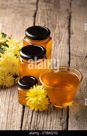 Miele biologico in vasetti di vetro a forma di favo e in una ciotola trasparente, cucchiaia di legno, fiori gialli su fondo di legno. Foto Stock