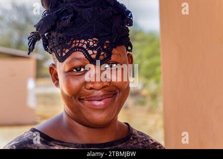 donna africana del villaggio, con trecce acconciature e copricapo, in piedi di fronte alla casa Foto Stock