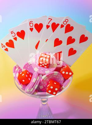 Vista dall'alto di Una mano di poker a filo rosso, chip da poker rosa e dadi rossi in un bicchiere da bere Martini su uno sfondo pastello luminoso e gradiente Foto Stock