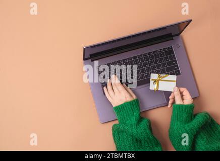 Concetto di shopping natalizio. Menzogna piatta. Mani da donna in un maglione verde sulla tastiera del computer portatile con carta regalo su sfondo rosso Foto Stock