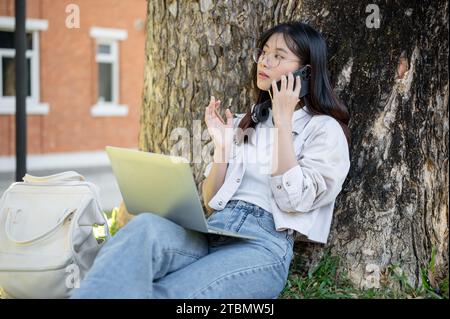 Una bellissima giovane studentessa asiatica sta parlando al telefono o chiamando qualcuno mentre lavora al suo computer portatile sotto l'albero in un parco universitario. Foto Stock