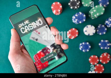 Mano con lo smartphone che mostra lo schermo con l'app di gioco del casinò online e le scommesse sui chip sul tavolo da gioco con sfondo verde feltro. Vista dall'alto. Foto Stock