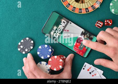 Giocatore di casinò online che accede all'app per giocare premendo la mano sullo schermo dello smartphone sul tavolo da gioco verde feltro con chip di scommesse e oggetto di gioco del casinò Foto Stock