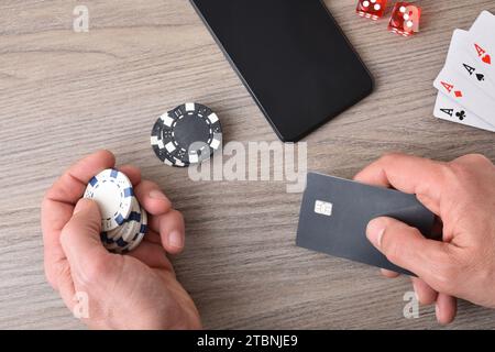 Giocatore di gioco del casinò che accede alla carta di pagamento per fare scommesse virtuali. Con patatine in mano e telefono e oggetti di gioco su un tavolo di legno. Vista dall'alto. Foto Stock