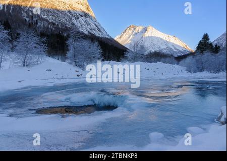 Una tranquilla mattinata d'inverno sulle montagne mostra un fiume parzialmente ghiacciato circondato da alberi ricoperti di neve e picchi aspri illuminati dalla luce del sole. Foto Stock