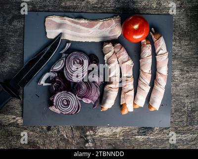 Piatto di salsicce ricoperte di pancetta, hot dog con cipolla di pomodoro e coltello da caccia, preparazione del cibo da picnic alla griglia all'aperto Foto Stock