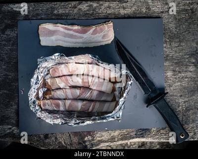 Piatto di salsicce ricoperte di pancetta, hot dog in stagno e coltello da caccia, preparazione del cibo da picnic alla griglia all'aperto Foto Stock