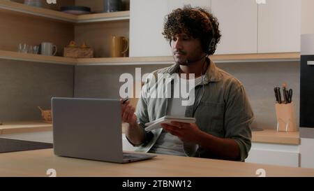 E-learning a distanza istruzione domestica uomo arabo in cuffia che guarda un webinar online di computer portatile in cucina studente indiano insegnante di Internet Foto Stock